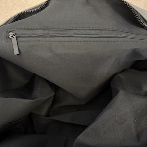 Puede incluir: El interior de un bolso gris oscuro que presenta un compartimento principal forrado con tela gris oscuro a juego. Un bolsillo horizontal con cremallera est&aacute; integrado en la parte superior de la pared interior, con un tirador de metal rectangular.