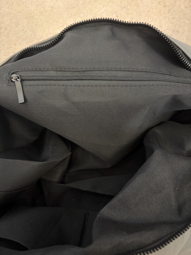 Puede incluir: El interior de un bolso gris oscuro que presenta un compartimento principal forrado con tela gris oscuro a juego. Un bolsillo horizontal con cremallera est&aacute; integrado en la parte superior de la pared interior, con un tirador de metal rectangular.