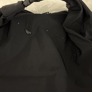 Puede incluir: Un bolso negro resistente al agua con cierre enrollable asegurado por una hebilla de pl&aacute;stico negra de liberaci&oacute;n lateral. La superficie de tela mate y lisa muestra varias gotas de agua peque&ntilde;as y claras sobre el panel frontal.