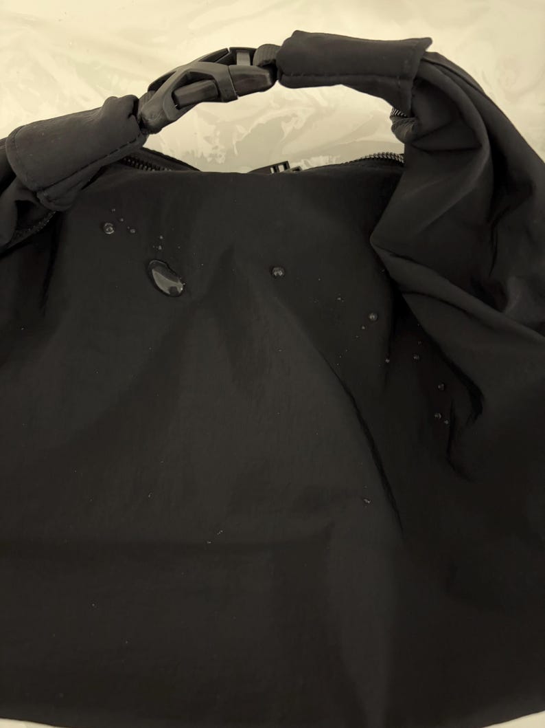 Puede incluir: Un bolso negro resistente al agua con cierre enrollable asegurado por una hebilla de pl&aacute;stico negra de liberaci&oacute;n lateral. La superficie de tela mate y lisa muestra varias gotas de agua peque&ntilde;as y claras sobre el panel frontal.