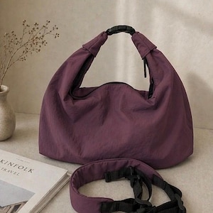 Puede incluir: Un bolso estilo hobo de color morado oscuro hecho de una tela suave y ligera. El bolso cuenta con una abertura superior asegurada por una hebilla de pl&aacute;stico negra. Se incluye una correa de hombro acolchada morada con cintas negras ajustables.