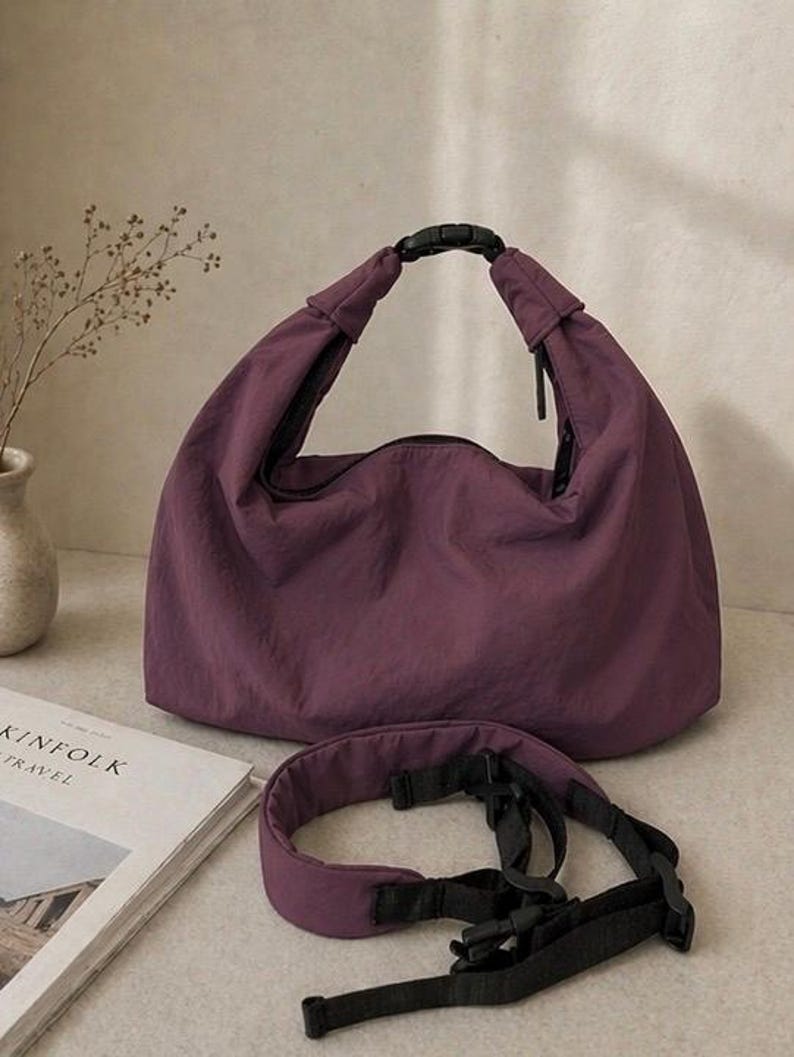 Puede incluir: Un bolso estilo hobo de color morado oscuro hecho de una tela suave y ligera. El bolso cuenta con una abertura superior asegurada por una hebilla de pl&aacute;stico negra. Se incluye una correa de hombro acolchada morada con cintas negras ajustables.