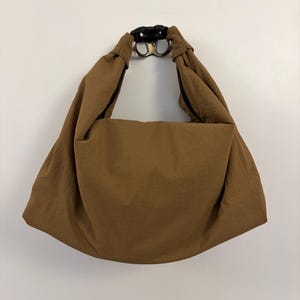 Puede incluir: Un bolso de estilo hobo marr&oacute;n hecho de una tela lisa de color s&oacute;lido. El bolso cuenta con una correa de hombro ancha e integrada que se conecta en la parte superior con una hebilla de pl&aacute;stico negra, creando una apariencia drapeada y fruncida.