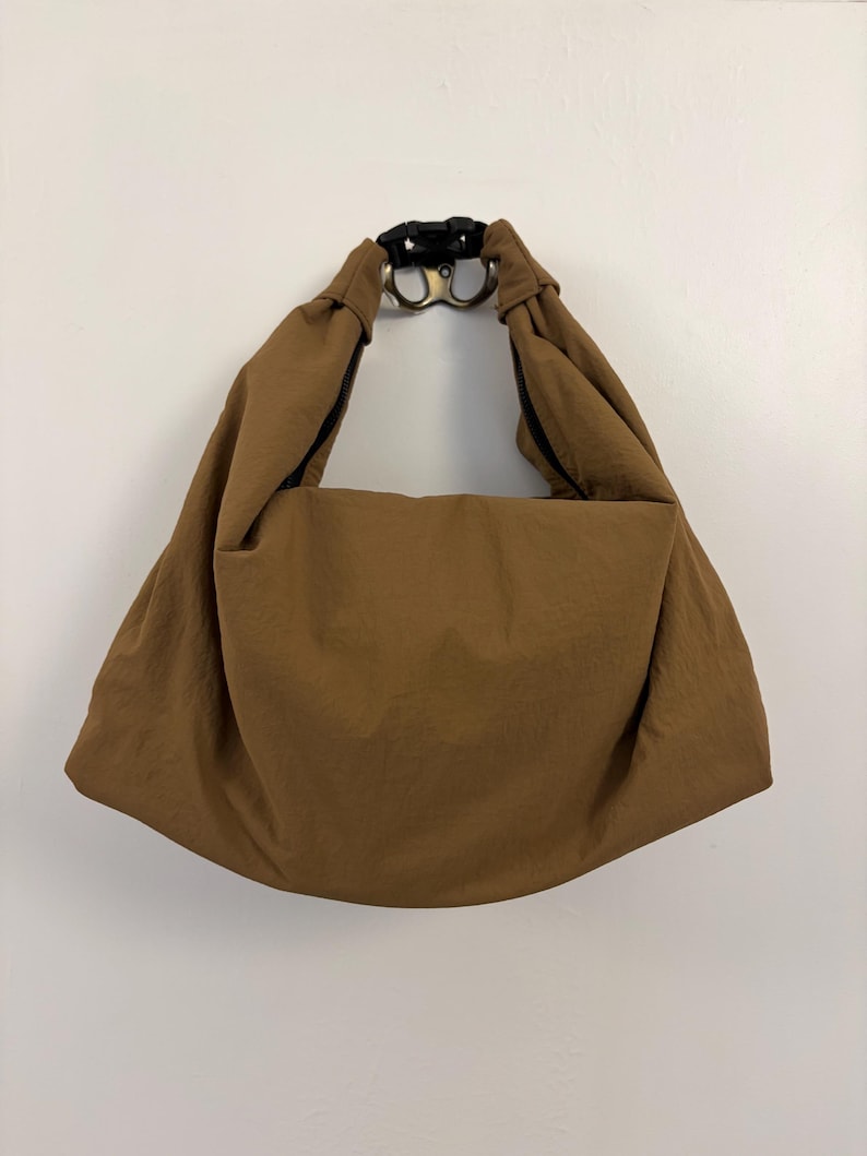 Puede incluir: Un bolso de estilo hobo marr&oacute;n hecho de una tela lisa de color s&oacute;lido. El bolso cuenta con una correa de hombro ancha e integrada que se conecta en la parte superior con una hebilla de pl&aacute;stico negra, creando una apariencia drapeada y fruncida.