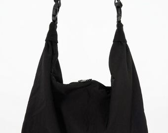Puede incluir: Bolso de hombro negro con correas ajustables y cierre de cremallera. El bolso está hecho de una tela ligera y texturizada. Las correas están unidas al bolso con hebillas de plástico negro. El bolso cuelga sobre un fondo blanco.