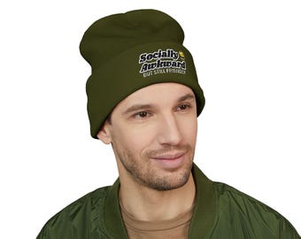 May include: Olive green beanie with the text "Socially Awkward But Still Friendly" in a white and yellow font. The beanie is worn by a person wearing a green bomber jacket.