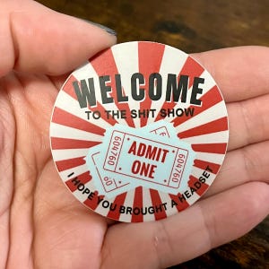 May include: A round, red and white striped sticker with the text "WELCOME TO THE SHIT SHOW" and a blue ticket that says "ADMIT ONE" with the text "I HOPE YOU BROUGHT A HEADSET" around the edge.