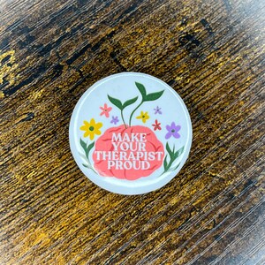 May include: A round, white button with the text "MAKE YOUR THERAPIST PROUD" in a stylized font. The design includes a pink brain with green leaves and colorful flowers.