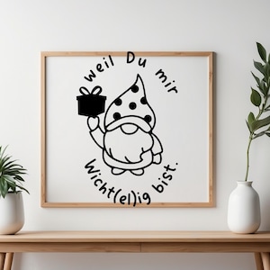 Può includere: Stampa incorniciata con un'illustrazione di gnomo che tiene un regalo. Lo gnomo indossa un cappello a pois e il testo recita "Weil Du mir wichtig bist." L'opera d'arte è esposta su un tavolo di legno con piante.