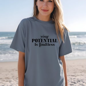 Könnte beinhalten: Hellblaues T-Shirt mit dem schwarzen Text "your POTENTIAL Is limitless". Das Shirt ist einfarbig und hat kurze Ärmel. Im Hintergrund ist ein Strand mit Meer und blauem Himmel.