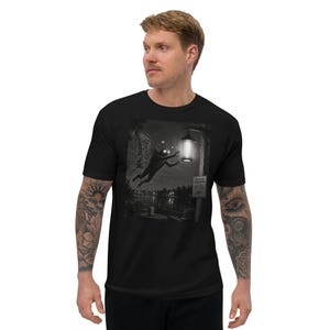 May include: A black t-shirt featuring a monochromatic graphic print of a moth-like creature with large wings flying toward a glowing street lamp near a lake. The scene includes a small sign with text and a table with chairs in the dark, detailed landscape.