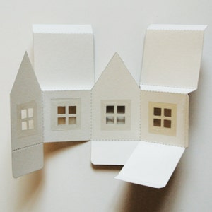 Puede incluir: Una casa de cart&oacute;n blanco con cuatro ventanas, cada una con un dise&ntilde;o diferente. La casa est&aacute; doblada y lista para ser ensamblada.