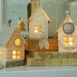 Puede incluir: Tres casas de papel en miniatura con ventanas iluminadas, decoradas con purpurina, pedrer&iacute;a y papel vintage. Las casas est&aacute;n dispuestas sobre una pila de libros antiguos.