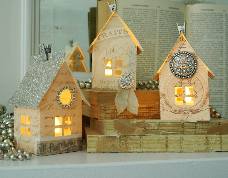 Puede incluir: Tres casas de papel en miniatura con ventanas iluminadas, decoradas con purpurina, pedrer&iacute;a y papel vintage. Las casas est&aacute;n dispuestas sobre una pila de libros antiguos.
