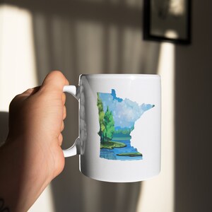 K&ouml;nnte beinhalten: Eine wei&szlig;e Keramik-Kaffeetasse mit einer bunten Illustration des Bundesstaates Minnesota, die einen See und B&auml;ume zeigt.