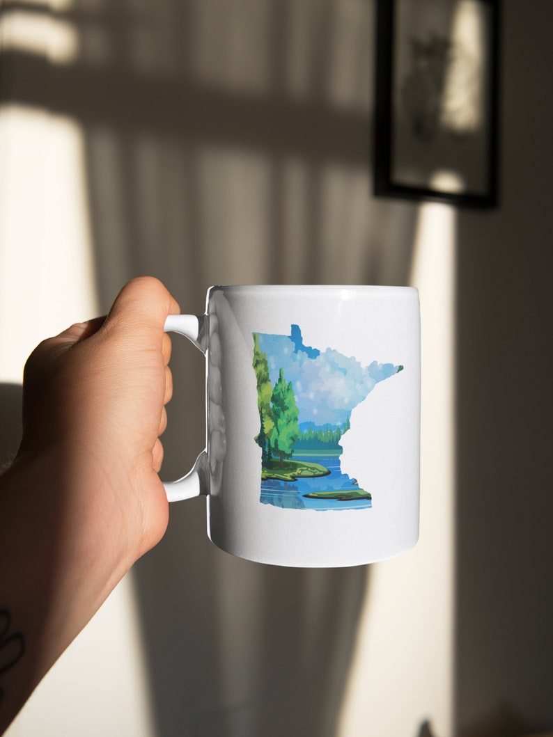 K&ouml;nnte beinhalten: Eine wei&szlig;e Keramik-Kaffeetasse mit einer bunten Illustration des Bundesstaates Minnesota, die einen See und B&auml;ume zeigt.