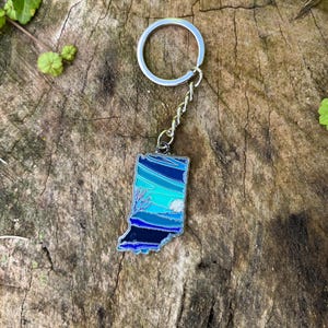 Puede incluir: Un llavero plateado con un contorno de esmalte azul y negro del estado de Indiana. El llavero tiene una cadena plateada y un anillo plateado.