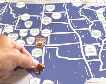 May include: A blue and white map of the United States with stickers of national parks. The stickers are placed on the map to show which parks have been visited. The stickers are round and have a picture of the park on them. The map is on a white surface.