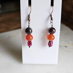 May include: A pair of dangle earrings with copper-colored hooks. Each earring features a dark gray bead, an orange bead, and a small, faceted, purple bead. The earrings are displayed on a white stand.