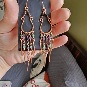 May include: Copper wire chandelier earrings with green, pink, and black beads. The earrings have a teardrop shape with dangling beaded strands and are held against a black card.