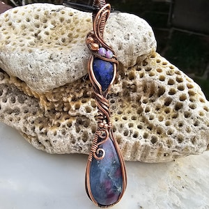 May include: A handcrafted pendant featuring two teardrop-shaped stones in shades of blue and purple, encased in copper wire. The wire is intricately woven, with small beads adding detail. The pendant hangs from a black cord, showcasing a unique, artistic design.