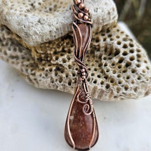 May include: A handcrafted pendant featuring a teardrop-shaped, reddish-brown stone encased in copper wire. The wire is intricately woven and coiled, with decorative beads at the top. The pendant hangs from a black cord.