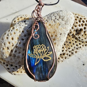 May include: A teardrop-shaped pendant with a copper wire frame and a black cord. The pendant features a blue and green stone with a gold lotus flower design. The wirework is intricate, and the pendant is displayed on a textured surface.