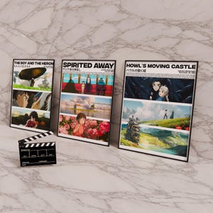 Pode incluir: Tr&ecirc;s cartazes de filmes emoldurados com ilustra&ccedil;&otilde;es de filmes do Studio Ghibli. Os cartazes s&atilde;o intitulados "O Menino e a Gar&ccedil;a", "A Viagem de Chihiro" e "O Castelo Animado".