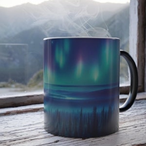 May include: A black coffee mug featuring a colourful aurora borealis design. The mug is placed on a white wooden surface with a window and mountains in the background.