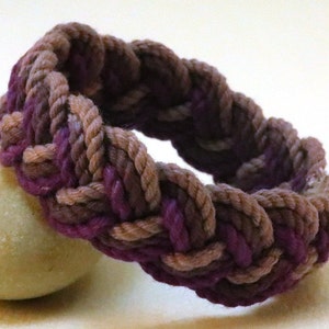 Puede incluir: Pulsera trenzada de cuerda en tonos morado, marrón y beige. La pulsera está hecha de cuerda gruesa y tejida, creando un accesorio texturizado y duradero. Los colores se mezclan, creando un diseño único. Una pulsera hecha a mano de inspiración náutica.