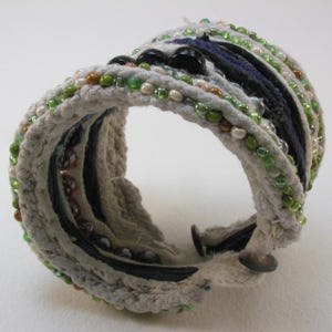 May include: A white crocheted bracelet with green, blue, and brown beads. The bracelet is made of multiple layers of rope and fabric.