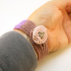 May include: A woven bracelet with a pink and purple striped band and a large, light pink, textured button.