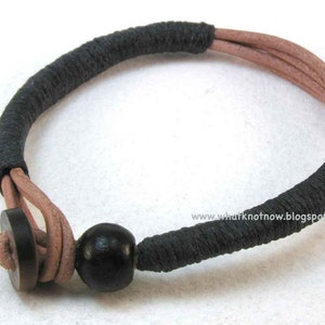 May include: Handmade bracelet featuring a combination of black wrapped sections and light brown leather strands. The bracelet has a wooden button clasp and a black wooden bead. A unique accessory.