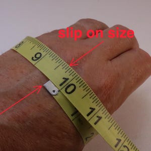 photo of hand with yellow cloth tape measure wrapped around the wide part showing how to measure your slip on size for fixed size bangles. A notation in red lettering points to the correct size on the tape measure.