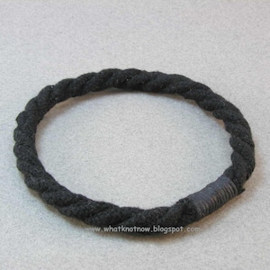 May include: A black braided rope bracelet with a black fabric covering the end of the rope.