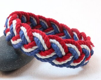Op de afbeelding: Een rood, wit en blauw gevlochten touw armband. Het touw is geweven in een patroon dat lijkt op een vlecht.