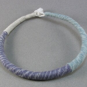 May include: A handmade necklace with a wrapped cord design. The necklace transitions from a light grey to a lavender and then to a light blue colour. The cord is knotted at the top, creating a circular shape.