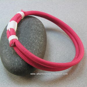 Op de afbeelding: Een felroze armband gemaakt van gevlochten touw, geaccentueerd met witte knopen. De armband is cirkelvormig en lijkt handgemaakt. De afbeelding is geplaatst tegen een grijze achtergrond, waardoor de kleur en het ontwerp van de armband worden benadrukt. De website www.whatknotnow.blogspot.com is zichtbaar.