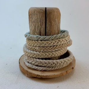 May include: A stack of four braided rope bracelets in shades of light blue and beige, displayed on a cylindrical piece of natural driftwood. The bracelets feature a woven texture and are arranged vertically around the wooden stand.