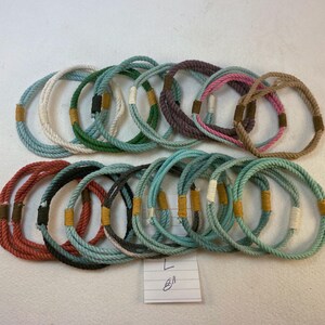 group of remainder string bracelets size large. A notation reading 8" is at the bottom of the frame  These bracelets are made with multiple strands of colored cotton cord bound with waxed thread whippings creating a fixed size slip on rope bracelet.