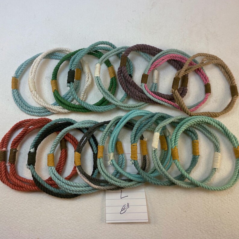 group of remainder string bracelets size large. A notation reading 8" is at the bottom of the frame  These bracelets are made with multiple strands of colored cotton cord bound with waxed thread whippings creating a fixed size slip on rope bracelet.
