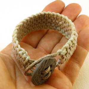 Op de afbeelding: Een beige gehaakt armband met een bruine knoopsluiting.