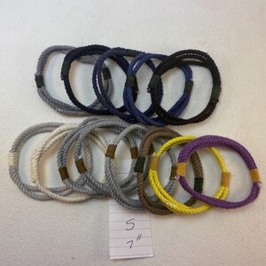 May include: Assortment of 12 colorful rope bracelets with brown leather accents. The bracelets are arranged in two rows. The text "5" and "7" is written on a white piece of paper.