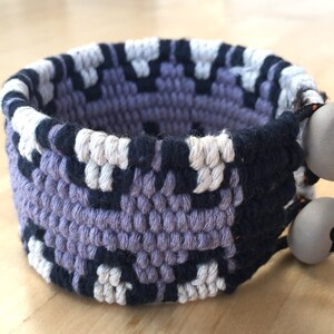 May include: A woven bracelet with a purple, black, and white pattern. The bracelet has two gray buttons with orange accents.