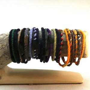 May include: A collection of colourful braided rope bracelets displayed on a wooden stand. The bracelets are made of different colours, including black, green, blue, purple, orange, and brown.