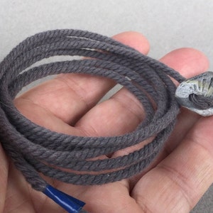 Puede incluir: Un cordón de algodón gris oscuro con una pequeña cuenta gris texturizada en forma de concha en el extremo.