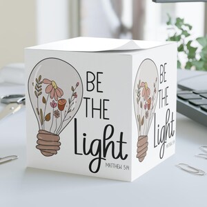 May include: A white cube-shaped notepad with the words "BE THE LIGHT" and a floral lightbulb design. The notepad has the Bible verse Matthew 5:14 printed on it. The design features soft pink, brown, and green colors.