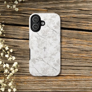 May include: A white phone case with a camouflage pattern of leaves and branches. A small cross is centered on the case. The case has a black border and a camera cutout.