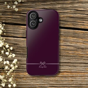 May include: A deep purple phone case with a bow graphic and the text "His way is better" in white. The case has a glossy finish and a dual-lens camera cutout. The phone case is on a wooden surface.