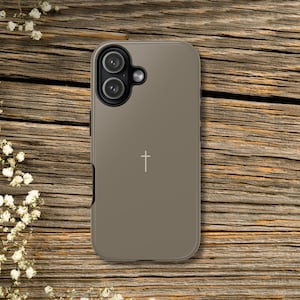 May include: A matte taupe smartphone case featuring a small, centered white cross icon. The case has a rectangular shape with rounded corners and a prominent circular camera cutout area in the upper left corner containing two camera lenses.